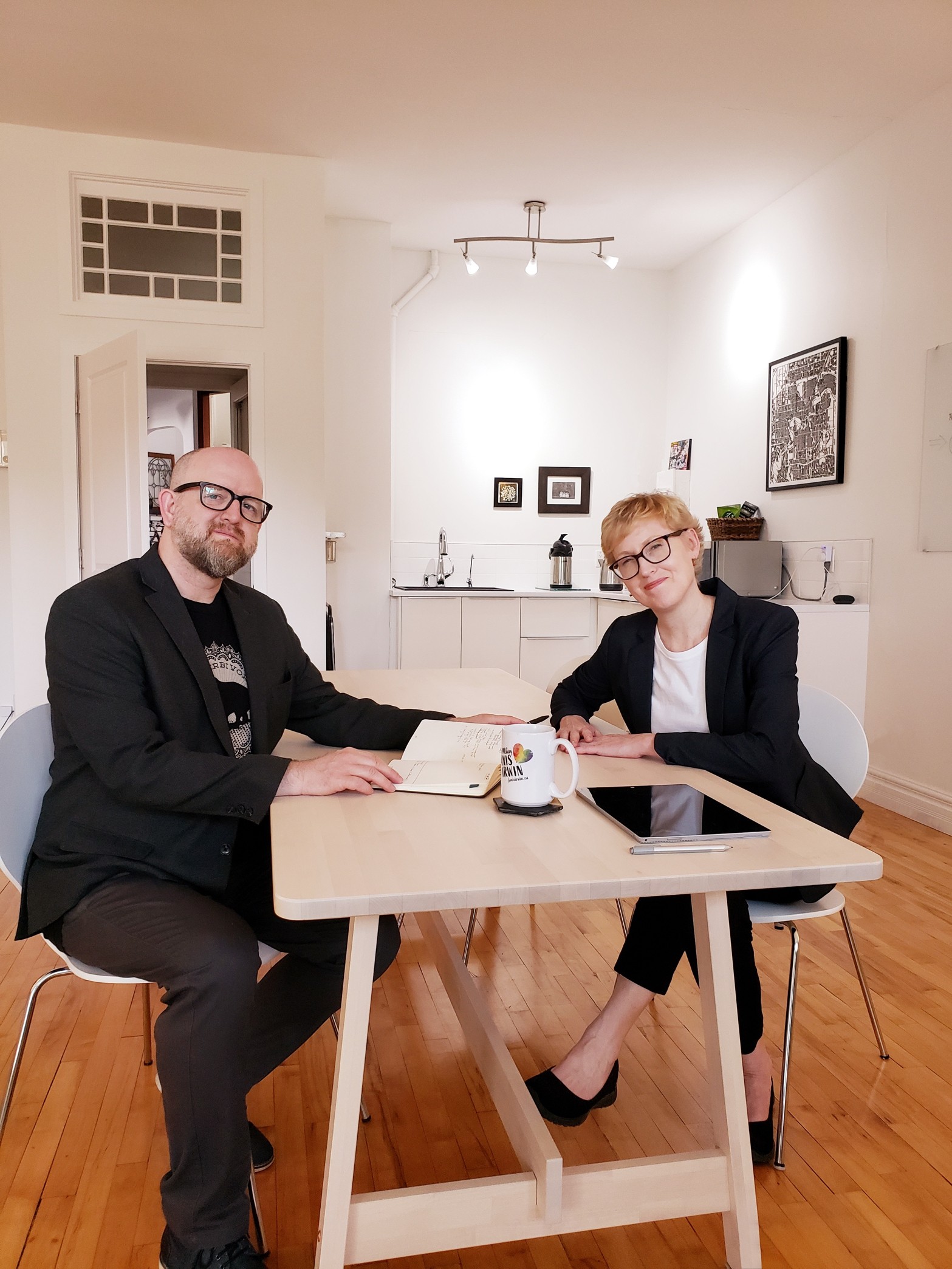 The two founders, Lara and Dwayne, sit opposite each other at a work table. Dwayne has a pen in one hand and the other is resting on an open journal. Lara has her palms down on the table with one hand slightly overlapping the other. They are smiling at the camera. Also on the table is a computer tablet with stylus and a coffee cup. The floor is natural hardwood. There is a door partway open in the background, with a transom window built into the wall above it. Also in the background is a kitchenette with a microwave and coffee carafe.