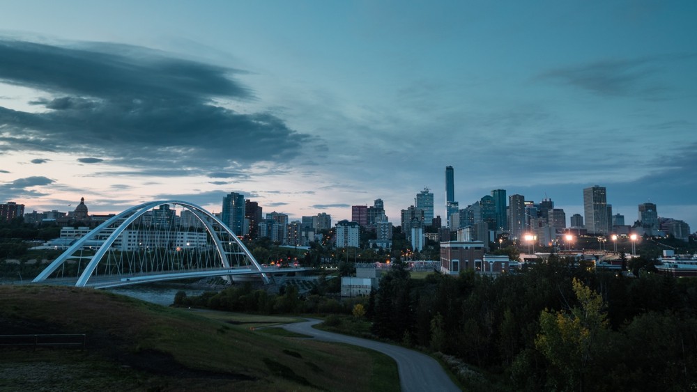 A wide view of the Edmonton skyline, looking from the south side of the North Saskatchewan River. The sun is setting in the west and there is a gentle sweep of clouds across the sky.